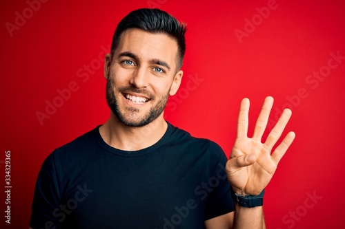 Fotomural Young handsome man wearing casual black t-shirt standing over isolated red background showing and pointing up with fingers number four while smiling confident and happy
