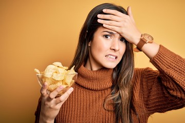 Sticker - Young beautiful girl holding bowl with chips potatoes standing over yellow background stressed with hand on head, shocked with shame and surprise face, angry and frustrated. Fear and upset