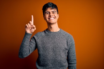 Young handsome man wearing casual sweater standing over isolated orange background showing and pointing up with finger number one while smiling confident and happy.