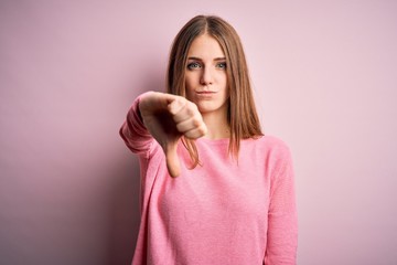 Wall Mural - Young beautiful redhead woman wearing casual sweater over isolated pink background looking unhappy and angry showing rejection and negative with thumbs down gesture. Bad expression.