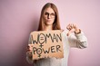 © Krakenimages.com - Young beautiful redhead woman asking for women rights holding banner over pink background with angry face, negative sign showing dislike with thumbs down, rejection concept