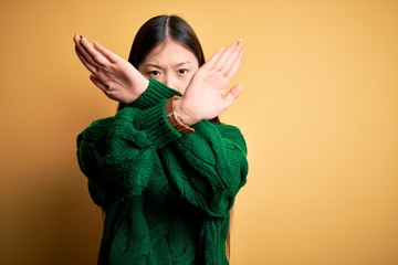 Poster - Young beautiful asian woman wearing green winter sweater over yellow isolated background Rejection expression crossing arms doing negative sign, angry face