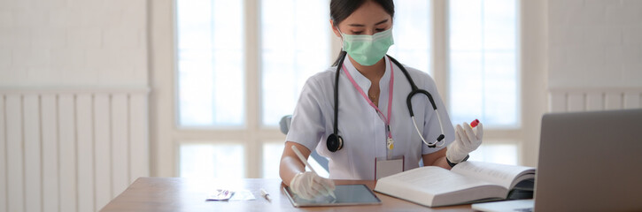 Wall Mural - Cropped shot of female doctor holding test tube and looking for Information in a Medical Book and laptop