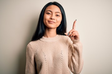 Young beautiful chinese woman wearing casual sweater over isolated white background showing and pointing up with finger number one while smiling confident and happy.