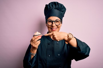 Middle age baker woman wearing cooker uniform and hat holding delicious chocolate cupcake very happy pointing with hand and finger