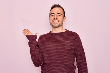 Young handsome man with blue eyes wearing casual sweater standing over pink background smiling cheerful presenting and pointing with palm of hand looking at the camera.