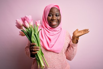 Canvas Print - Young african american plus size woman wearing muslim hijab holding bouquet of pink tulips smiling cheerful presenting and pointing with palm of hand looking at the camera.