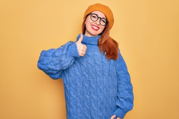 Wall Mural - Young beautiful redhead woman wearing french beret and glasses over yellow background doing happy thumbs up gesture with hand. Approving expression looking at the camera showing success.