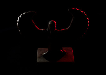 Closeup to a Isis. female old winged egyptian god mini figurine into darkness iluminated with white and red lights over black background 