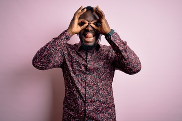 Poster - Young handsome african american man wearing casual shirt standing over pink background doing ok gesture like binoculars sticking tongue out, eyes looking through fingers. Crazy expression.