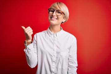 Wall Mural - Young blonde business woman with short hair wearing glasses over red background smiling with happy face looking and pointing to the side with thumb up.