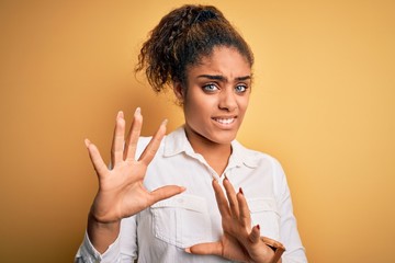 Sticker - Young beautiful african american girl wearing casual shirt standing over yellow background disgusted expression, displeased and fearful doing disgust face because aversion reaction. With hands raised