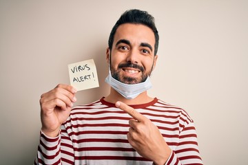 Poster - Young handsome man with beard wearing medical mask holding virus alert reminder very happy pointing with hand and finger