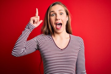 Wall Mural - Young beautiful blonde woman wearing casual striped t-shirt over isolated red background pointing finger up with successful idea. Exited and happy. Number one.