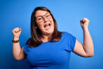 Beautiful brunette plus size woman wearing casual t-shirt over isolated blue background celebrating surprised and amazed for success with arms raised and open eyes. Winner concept.