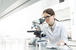 © pressmaster - Serious female researcher in whitecoat looking in microscope by workplace