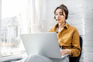 Wall Mural - Young woman with a headset working online on computer while sitting on the window sill at home. Concept of studying or working from home online