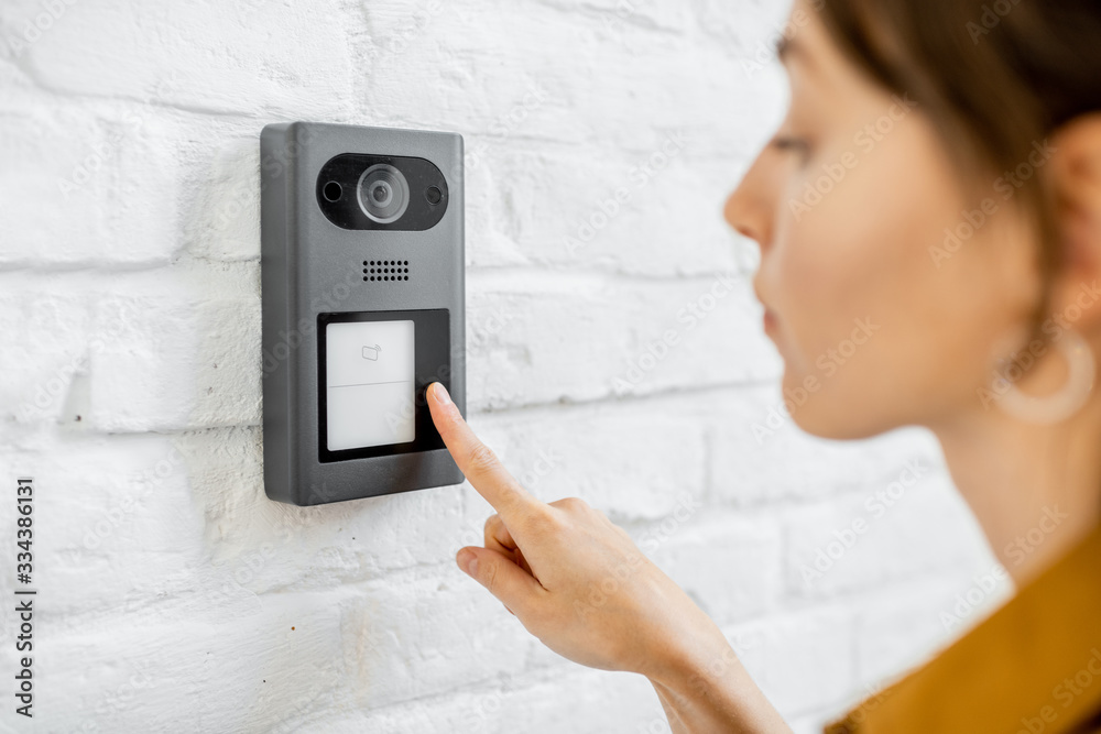 Woman rings the house intercom with a camera installed on the white brick wall - obrazy, fototapety, plakaty 