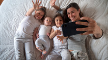 Authentic Shot Of Happy Mother With Her Kids Are Making A Selfie Or Video Call To Father Or Relatives In A Bed. Concept Of Technology, New Generation,family, Connection, Parenthood, Authenticity