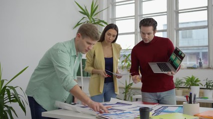 Wall Mural - Tracking medium shot of young startup team of three specialists standing around table in office and discussing website templates presented on laptop and printed on papers