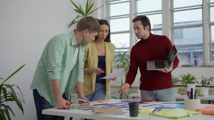 Wall Mural - Young startup team of three standing around table in office and discussing application design templates presented on laptop and printed on papers