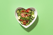 Fresh Salad With Tomato, Cucumber, Vegetables, Microgreen Radishes In Plate Shape Of Heart On Green. View From Above. Concept Vegan And Healthy Eating.