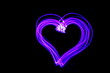 Long exposure photograph of neon blue and purple streaks of light in a heart shape outline symbol, parallel lines pattern against a black background. Light painting photography.