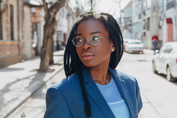 Wall Mural - Close up portrait of a beautiful young african american woman with pigtail hairstyle in a blue jacket and glasses smiling and walking along the street