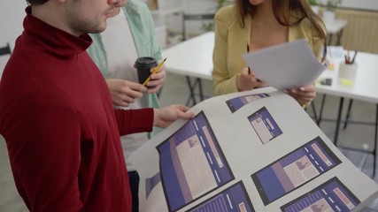 Wall Mural - Tilt down medium shot of young startup team of three business people discussing printout with web application templates for different devices standing in office
