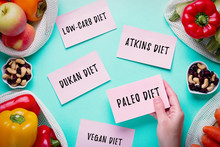 Woman Choosing Between Popular Diets For Weight Loss With Fresh Fruits, Nuts And Vegetables In Background. Stickers With Popular Diet Names On Blue, Healthy Lifestyle Concept, Flat Lay, Top View