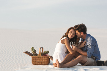 Wall Mural - young couple hugging while sitting on blanket with basket of fruits and acoustic guitar on beach