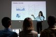 © pressmaster - Young woman pointing at projection screen with graph variations and giving speech on own startup project in front of investors