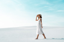 Side View Of Smiling Beautiful Girl In White Dress And Straw Hat Walking On Sandy Beach With Blue Sky