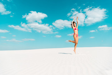 Wall Mural - happy beautiful sexy girl in swimsuit jumping with hands in air on sandy beach with blue sky and clouds