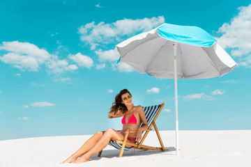 Wall Mural - smiling beautiful sexy girl in swimsuit and sunglasses sitting in deck chair under umbrella on sandy beach