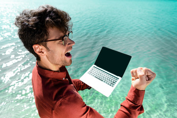 Wall Mural - excited handsome freelancer with laptop standing in turquoise water