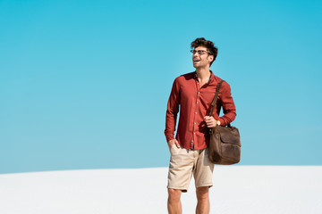 Wall Mural - smiling handsome man with leather bag on sandy beach against clear blue sky