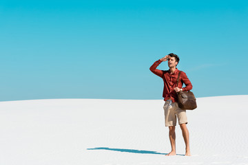 Wall Mural - handsome man with leather bag looking away on sandy beach against clear blue sky