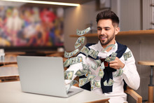 Man With Modern Laptop And Flying Dollar Banknotes At Table Indoors. People Make Money Online
