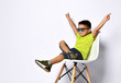 Kid in sunglasses, yellow t-shirt, denim shorts, khaki sneakers. Smiling showing victory sign, sitting on chair. Isolated on white