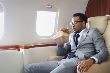 Wall Mural - Worried african american businessman breathing with paper bag while having panic attack during flight on private plane