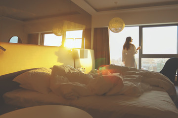 Silhouette of a young beautiful woman with cup of coffee in the white bathrobe on the hotel window background at the yellow sunrise time