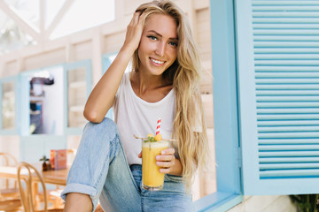 Inspired woman with long hairstyle holding glass of juice and posing with smile. Portrait of tanned blonde model in jeans sitting on sill and drinking cocktail.