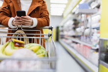 Woman With Smart Phone Pushing Shopping Cart In Supermarket