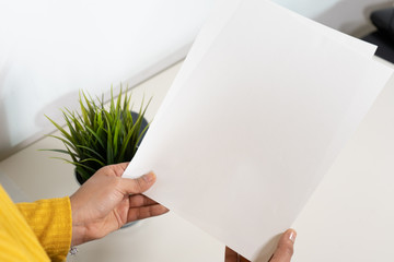 Close up of a set of two papers hold by a caucasian woman with yellow shirt and bracelet sitting on a desk with a plant