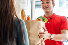 Asian Deliver Man Worker In Red Color Uniform Handling Bag Of Food, Fruit, Vegetable Give To Young Beautiful Female Costumer In Front Of The House. Postman And Express Grocery Delivery Service Concept