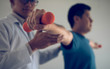 © wutzkoh - Asian physiotherapist helping a patient lifting dumbbells work through his recovery with weights in clinic room.