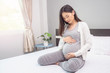 © Have a nice day  - beautiful asian pregnant woman smiling and hands rubbing firmly on her baby stomach, joyfully and happiness, sitting on the white bed relaxing and resting in the bedroom with light shining from window
