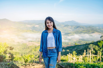 Beautiful asian tourist traveler girl on holiday adventure traveling on the mountains, walking along the path smiling in the morning sunrise and misty mountains view wearing a denon jacket and jeans