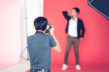 Back View Of Male Asian Professional Photographer Taking Picture Of Male Model In Red Isolated Background With Studio Lighting Umbrella Soft Box, Representing Photographer Studio Lighting Concept